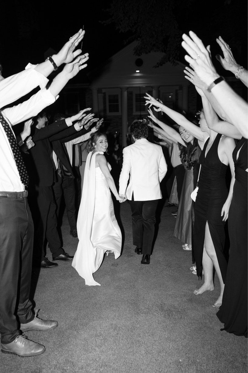 Wedding guests form a tunnel with raised hands as the couple makes their grand exit in black and white.