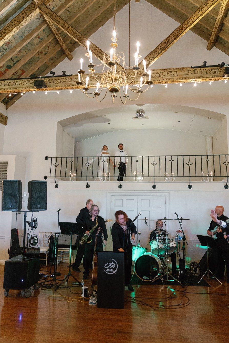 A live band performs in an elegant ballroom with exposed wood beams and a grand chandelier.