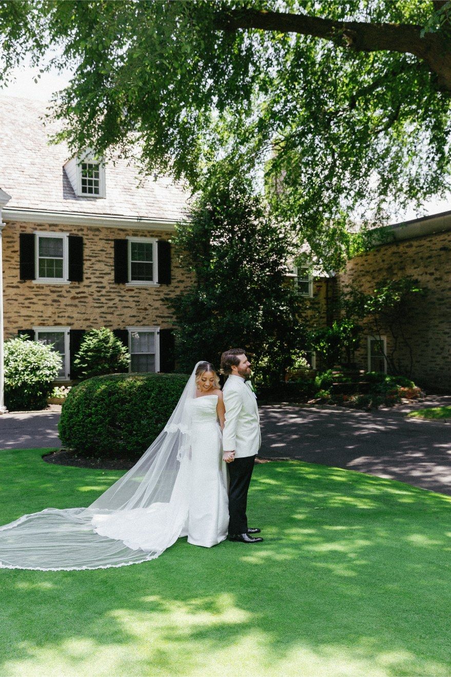 A wedding couple shares an intimate moment on a lush green lawn outside a stone house with white trim and hanging trees.
