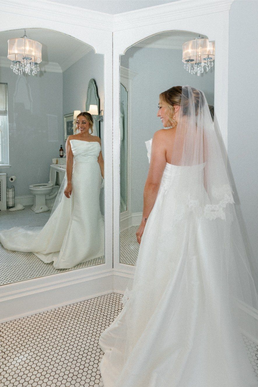 Elegant wedding gown with long train and veil reflected in bathroom mirror with crystal chandelier lighting.