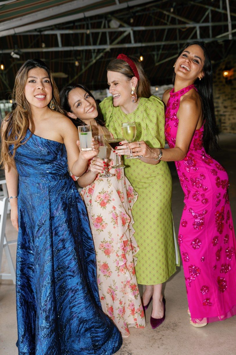 Four friends in colorful formal dresses smile and raise wine glasses for a toast at an indoor celebration.