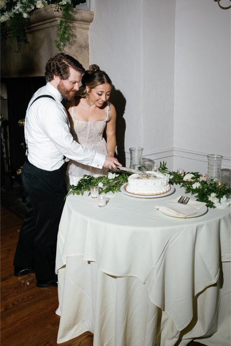 A couple shares a romantic moment while cutting their wedding cake at a white-clothed reception table decorated with greenery.