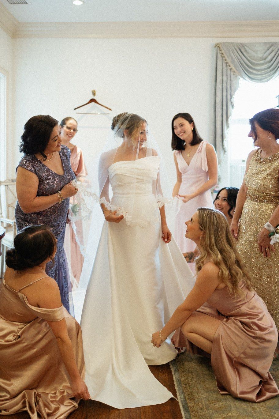 A wedding party helps the bride get ready in a room with elegant white dresses and gold detailed gowns.