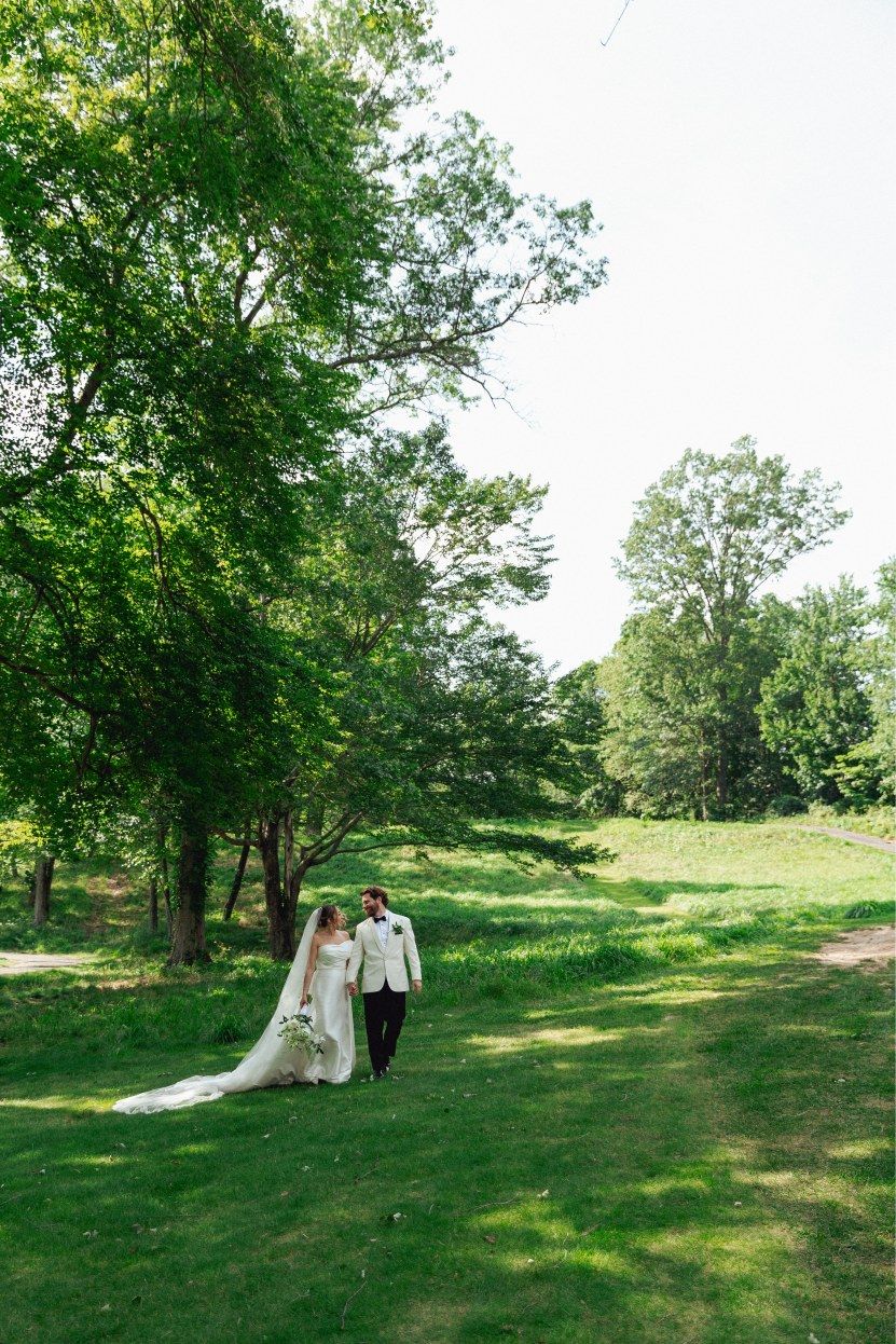 A bride and groom walk hand in hand through a sunlit garden with trees and rolling hills in the background.