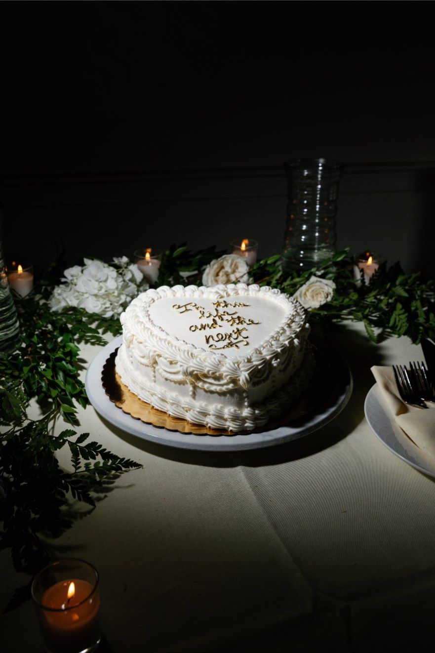 A romantic heart-shaped cake with white frosting displayed on a table with greenery and soft candlelight.