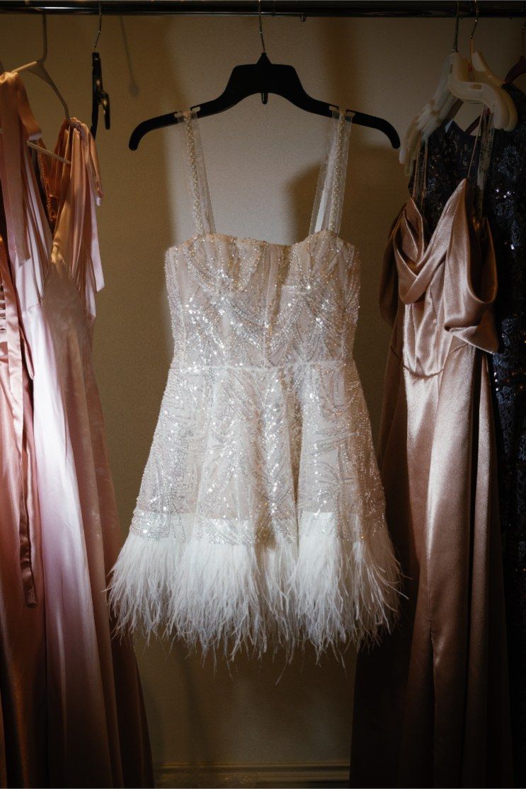 White lace mini dress with feather hem detail and corset bodice hanging on black hanger among other formal dresses.
