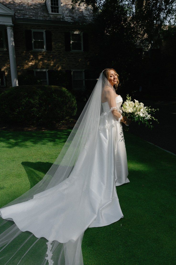 A bride in a flowing white wedding gown with a dramatic train stands on a green lawn at dusk.