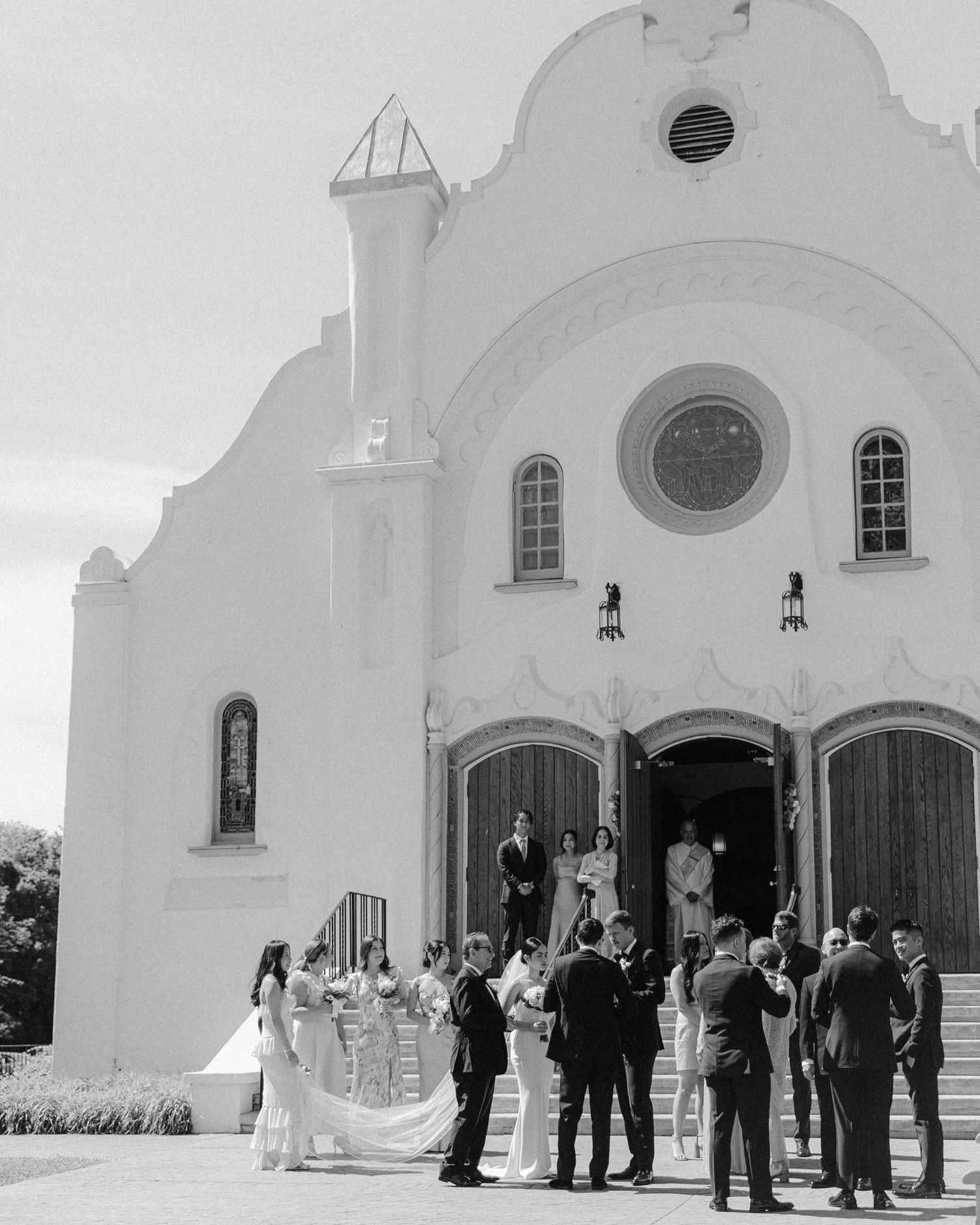 People gather outside a white Spanish Mission style church with arched doorways during what appears to be a wedding ceremony.