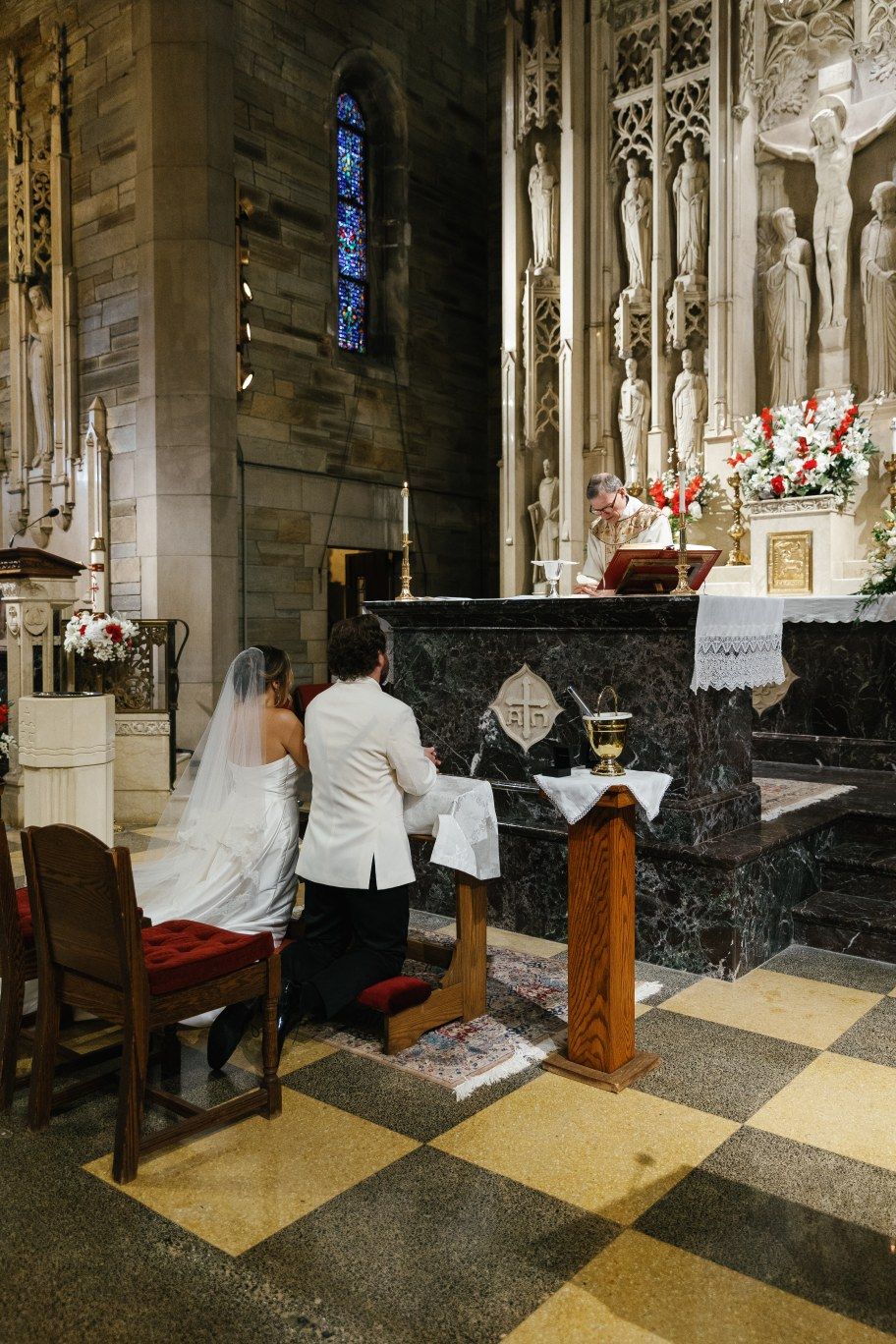 A wedding couple kneels at a marble altar inside a gothic cathedral with stained glass windows and ornate stone carvings.