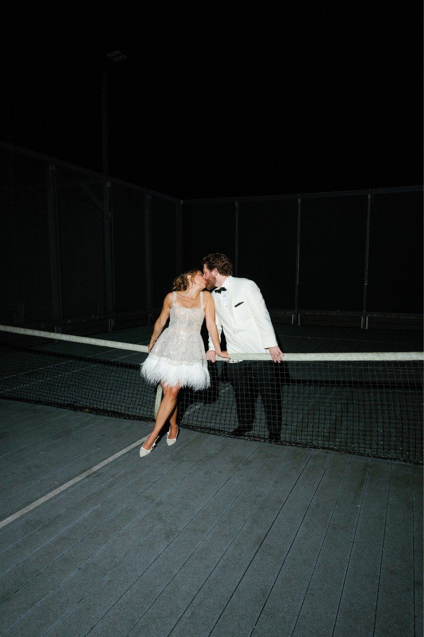 A romantic nighttime portrait of newlyweds sharing an intimate moment by a tennis net.
