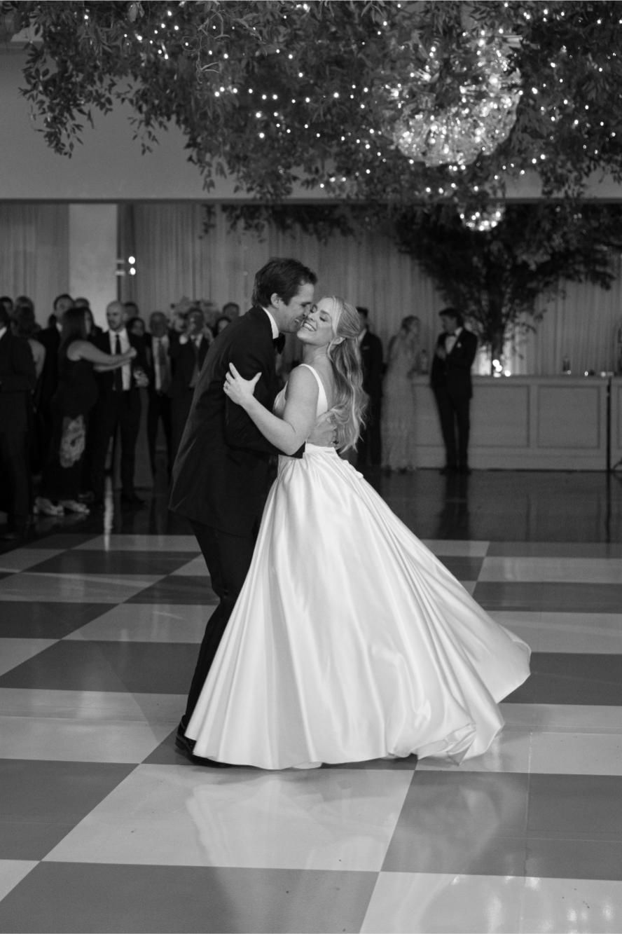 A romantic black and white wedding photograph of a couple dancing on a checkered ballroom floor under twinkling lights.