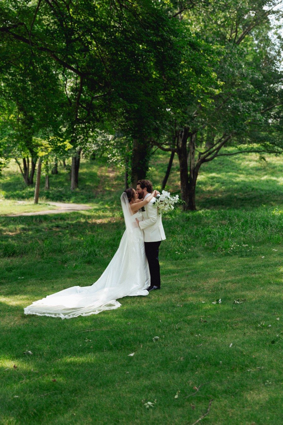 A newlywed couple shares an intimate moment on a lush green lawn surrounded by trees on their wedding day.