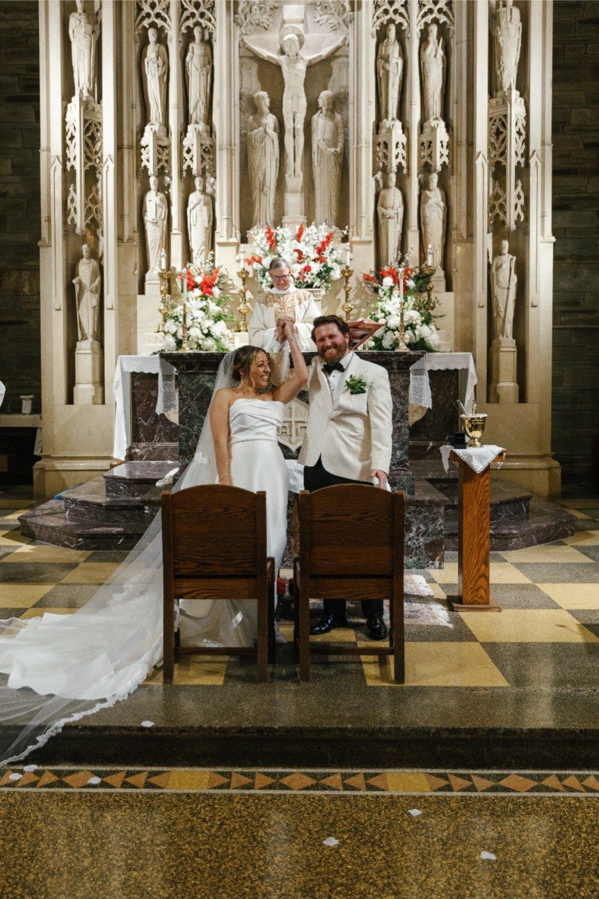 Newlyweds sit together at a Catholic church altar with ornate Gothic architecture and floral arrangements.