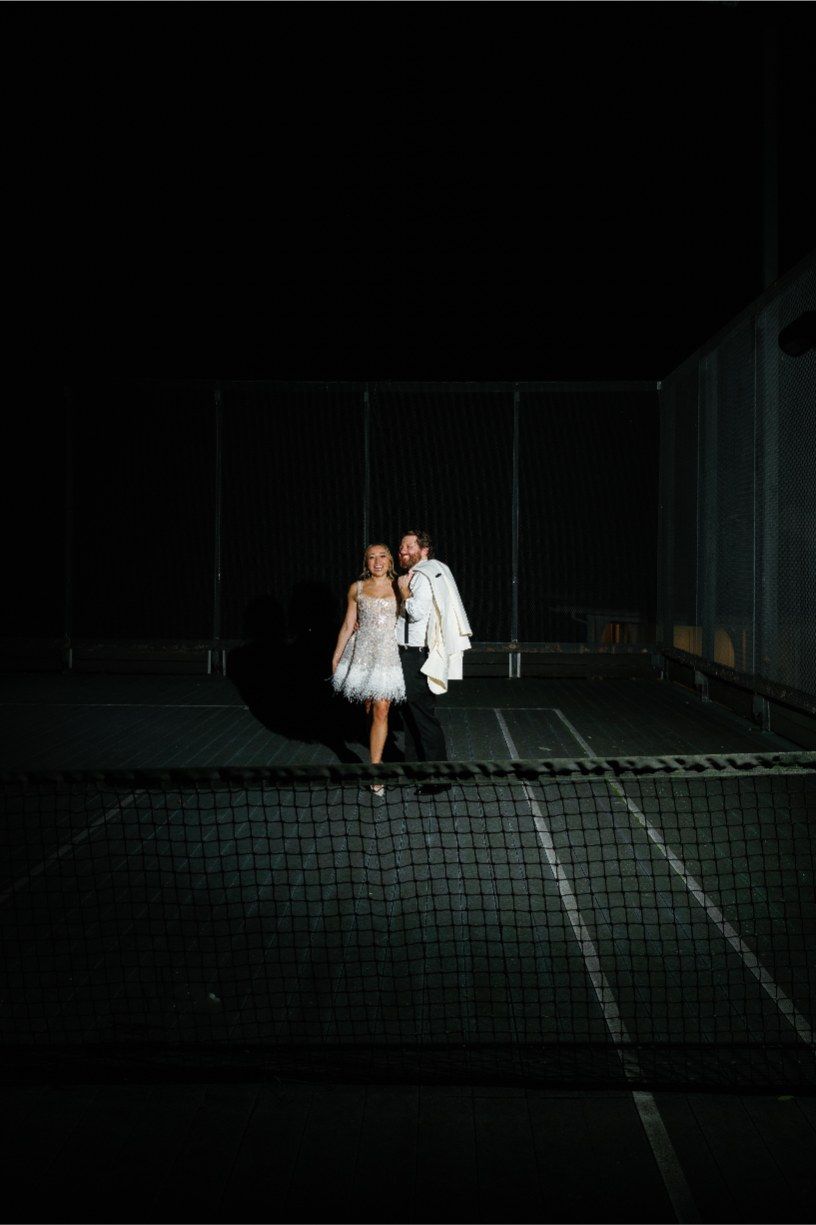 A couple in formal attire stands on darkened tennis courts at night creating a dramatic silhouette effect.