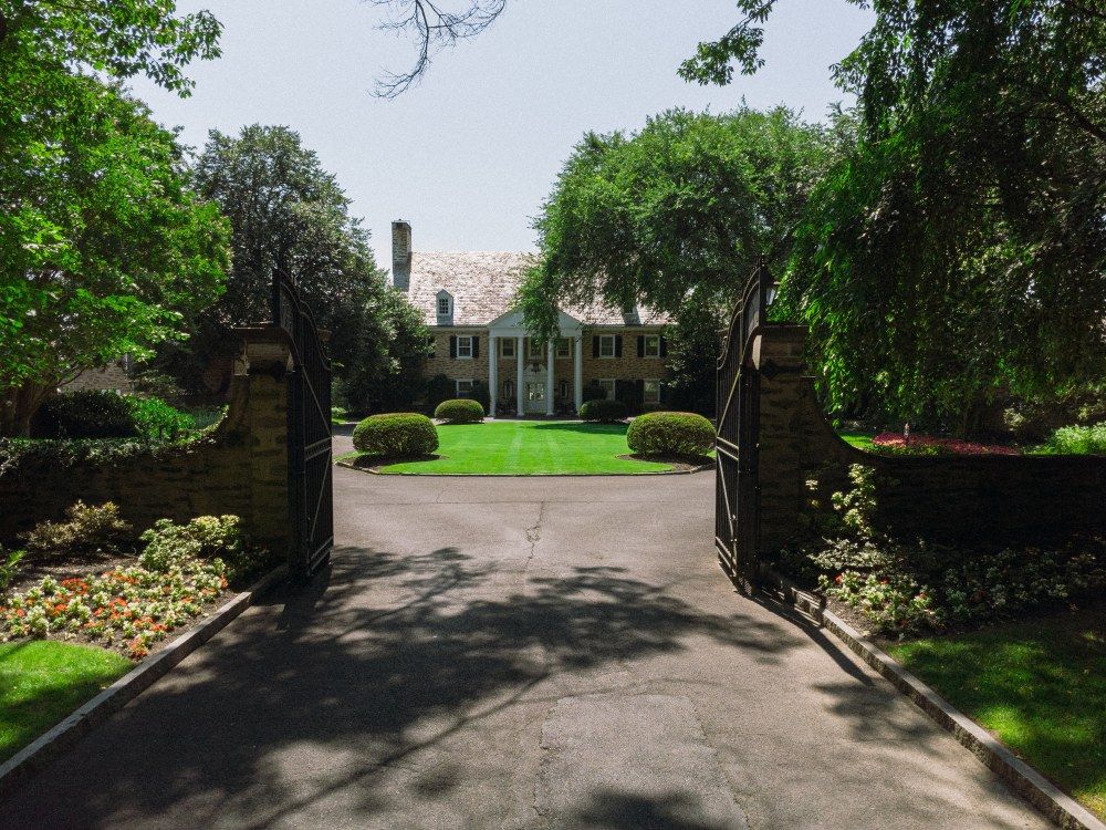 A winding suburban driveway entrance flanked by lush greenery and mature trees on a sunny day.