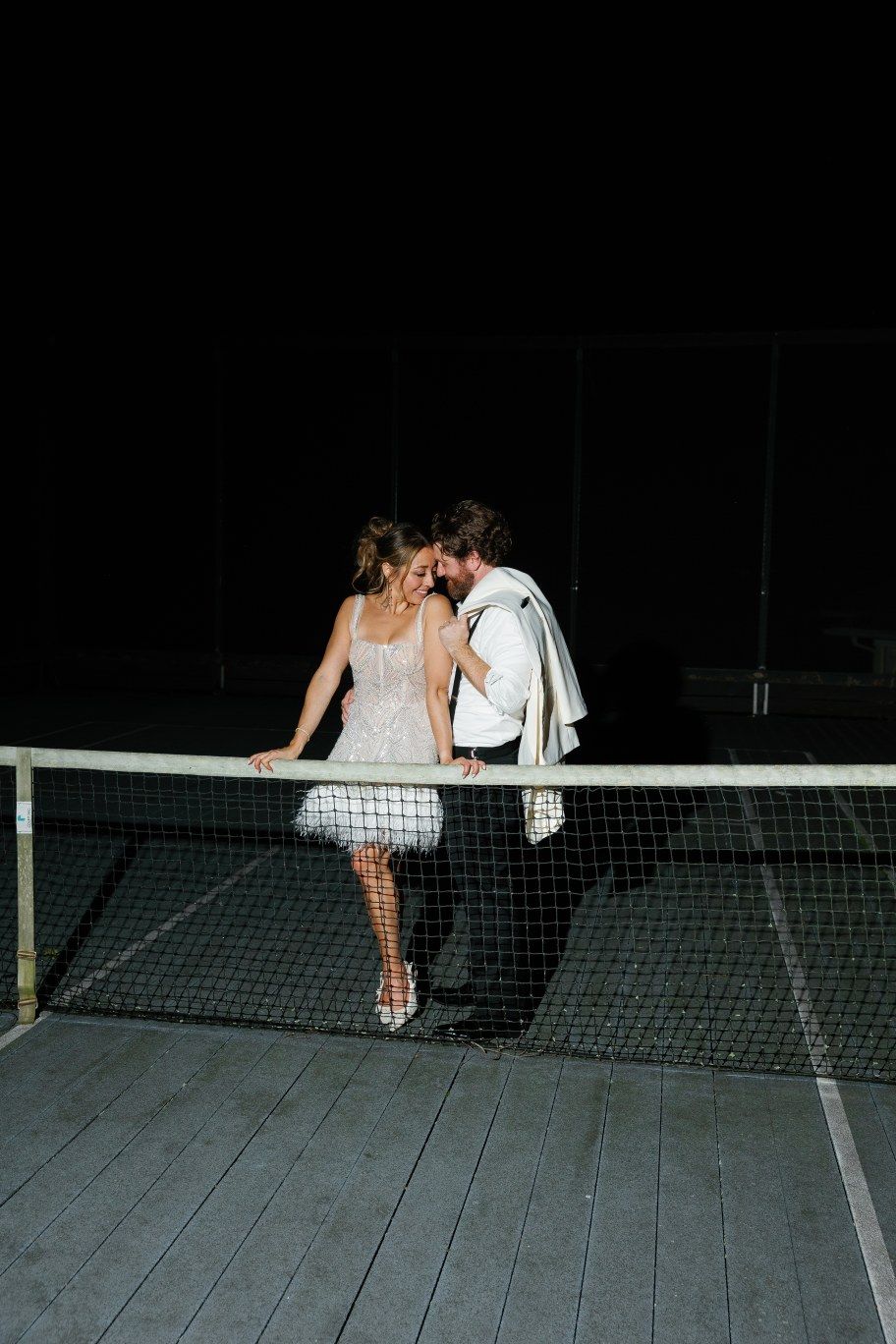 Two figures in white dresses lean dramatically across a volleyball net at night creating a mirrored pose.