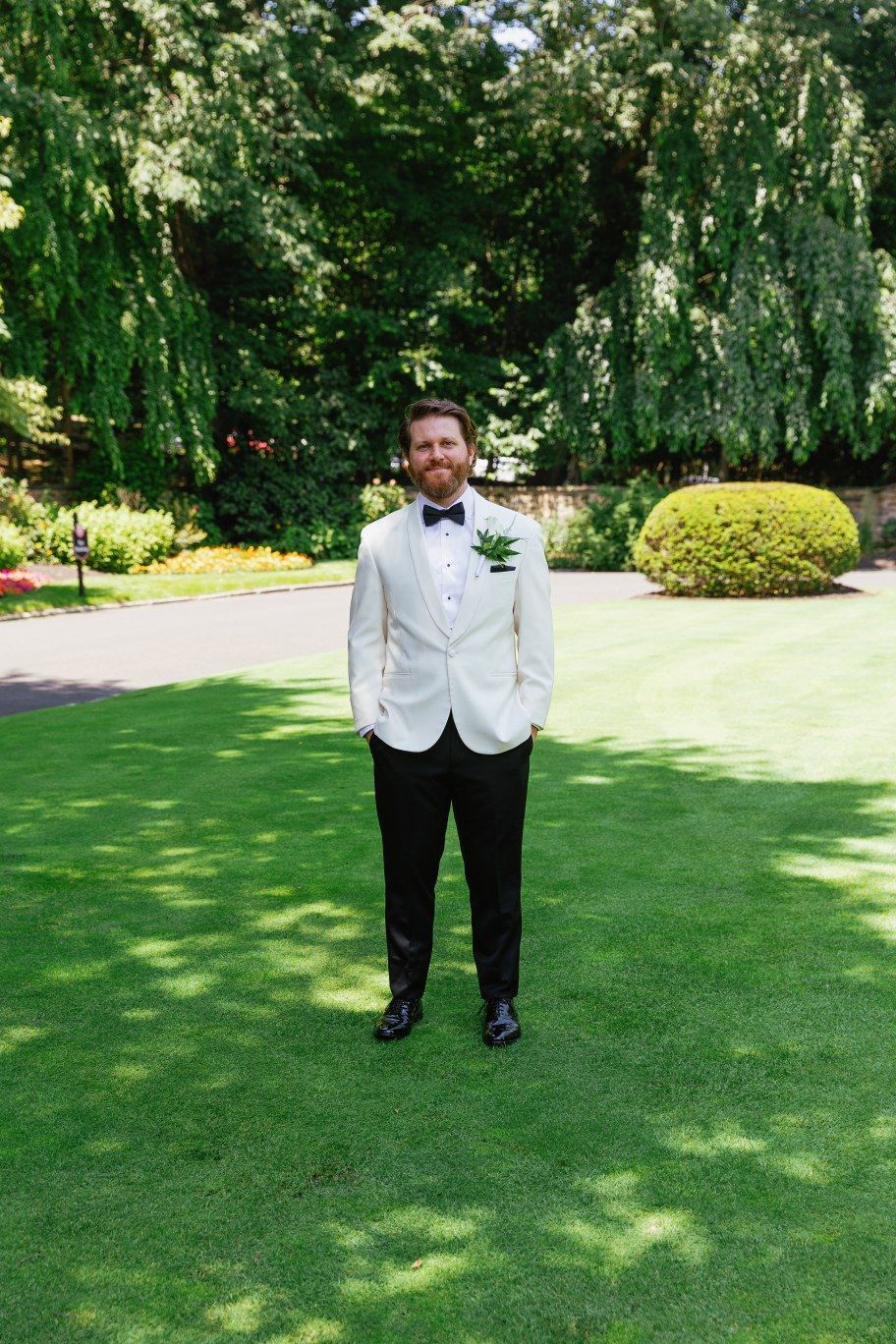 Someone wearing a white dinner jacket and black bow tie stands on a manicured lawn with weeping willows in the background.