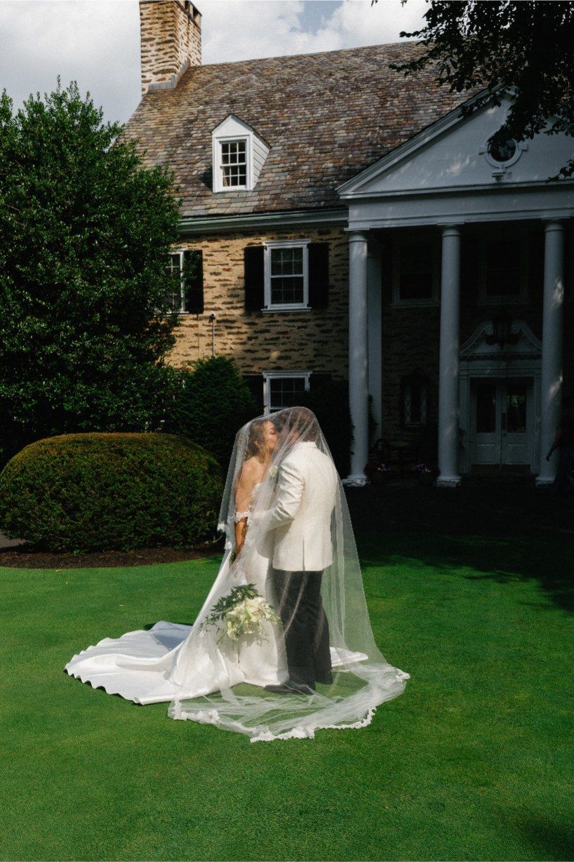 Wedding couple embracing on a lush green lawn in front of a classic stone house with white columns.