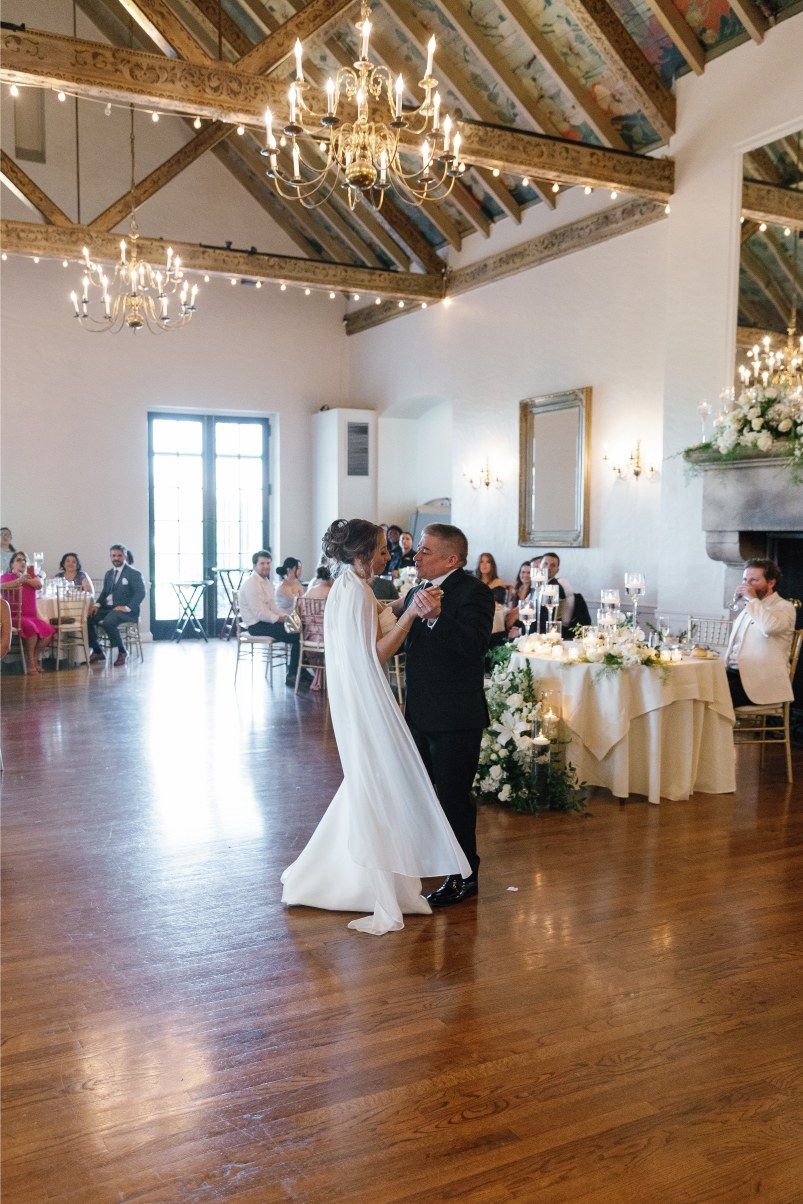 A couple shares their first dance at an elegant reception venue with exposed wood beams and crystal chandeliers.