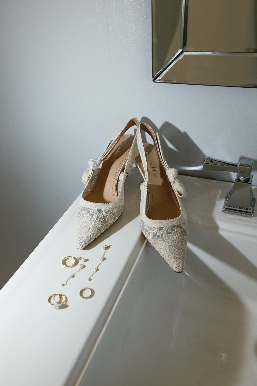 Elegant white lace wedding heels on a white surface with wedding rings displayed next to them.
