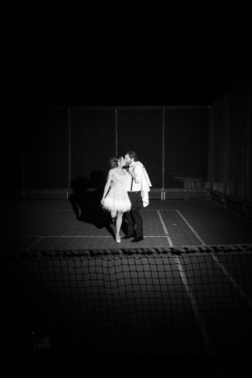 Black and white aerial view of a figure in white dress playing on a rooftop sports court at night.