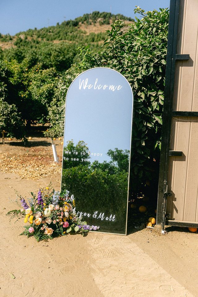Ornate welcome mirror with floral arrangement displayed against vineyard backdrop at outdoor wedding venue.