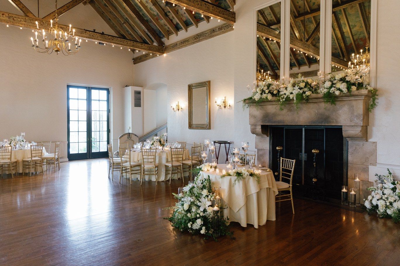 Elegant wedding reception venue with chandelier, hardwood floors and white floral decorations in a rustic-chic setting.