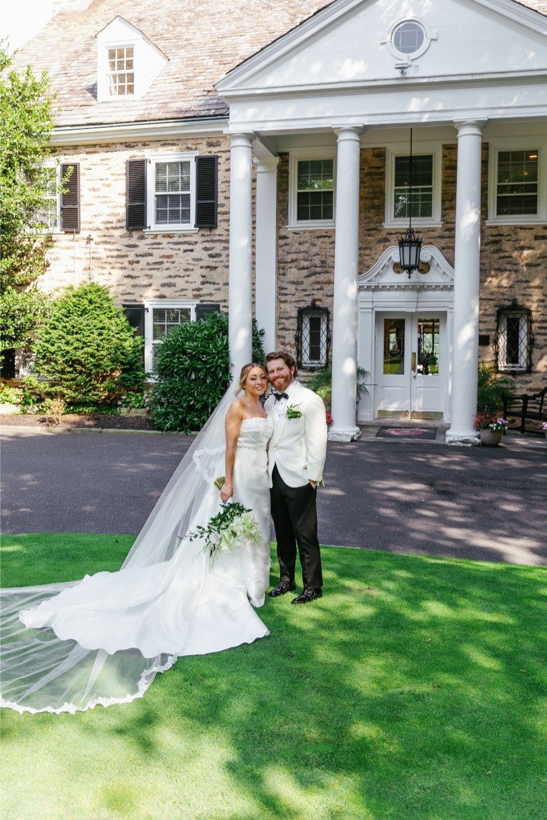 A couple in formal attire stand under a grand portico of an elegant mansion during a wedding photo shoot.