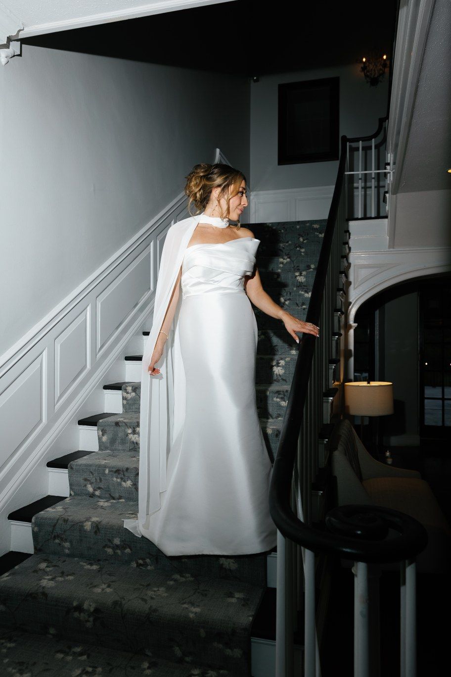 A person in a flowing white gown descends a carpeted staircase in a dimly lit interior setting with elegant grey walls.