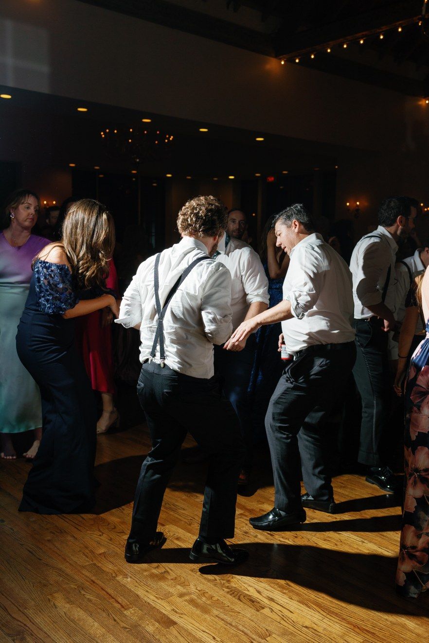 Wedding guests dance together in a dimly lit venue on a wooden dance floor during a reception celebration.