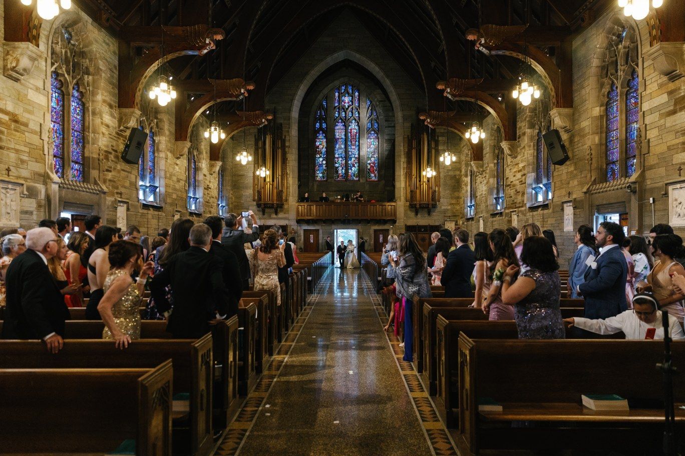 Interior view of a grand church during a ceremony with guests seated in wooden pews beneath Gothic arches and stained glass.