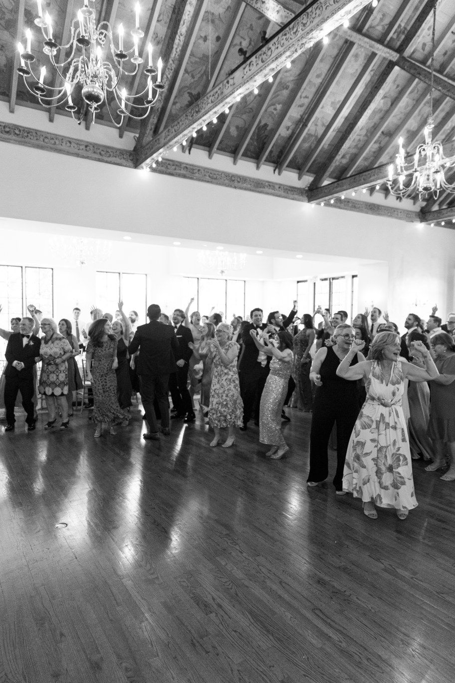 Wedding guests line up in formation on a wooden dance floor under chandeliers.