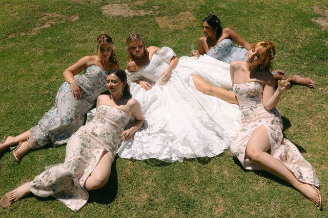 Group in matching dresses laying in a star formation on grass during a wedding photo shoot.