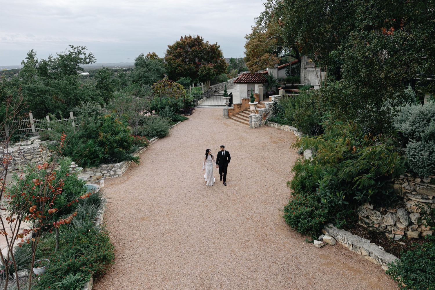 A winding gravel path curves through a lush garden with trees, shrubs and stone walls on both sides.