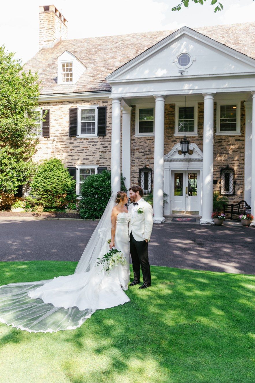 A couple poses for a wedding photo in front of a grand stone mansion with white columns and lush landscaping.