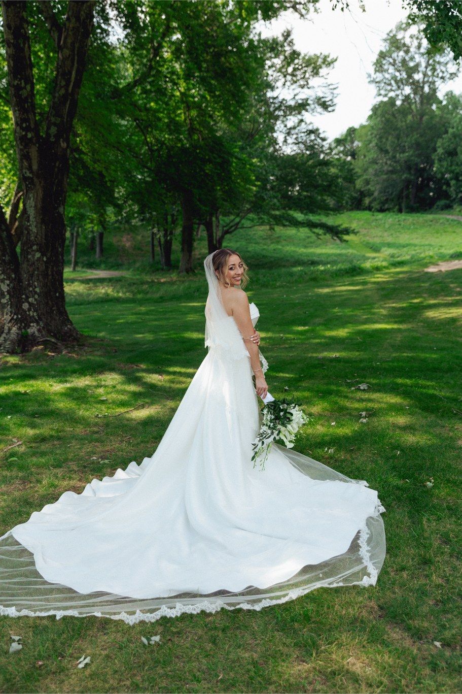 Elegant bride in a flowing white gown with long train poses on grass in a serene garden setting.