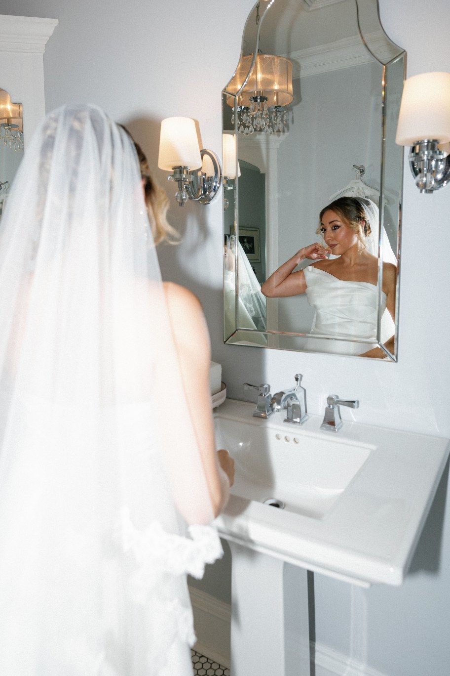 A bride in a white wedding gown and veil looks at her reflection in an ornate bathroom mirror with crystal sconces.