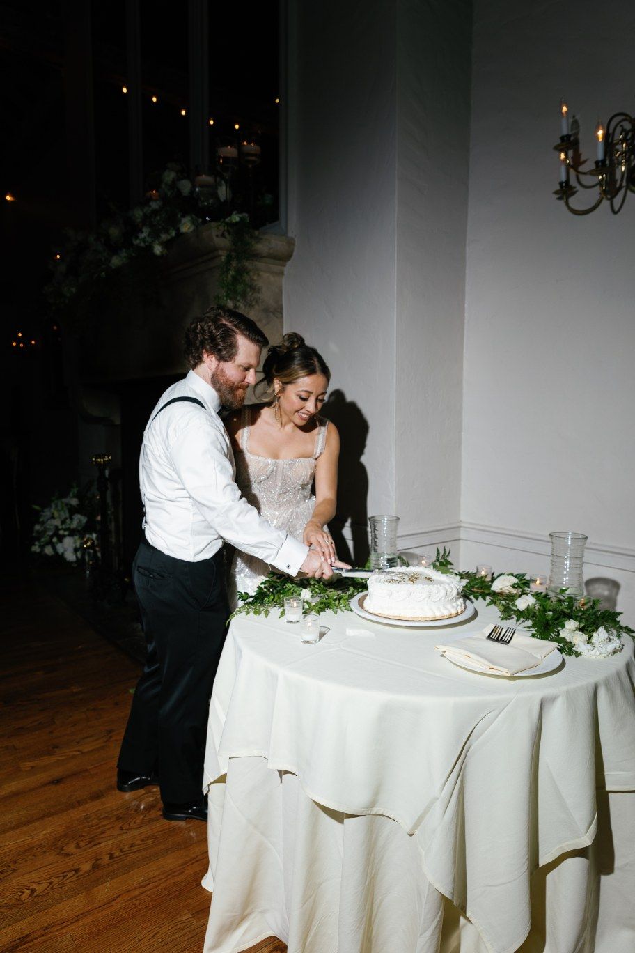 A couple cuts their wedding cake together at a reception table decorated with white linens and greenery.
