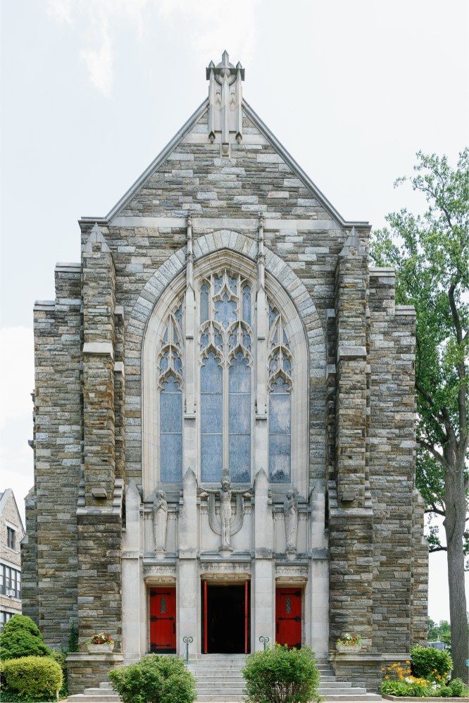 Gothic stone church facade with pointed arch stained glass window, red doors, and ornate architectural details.