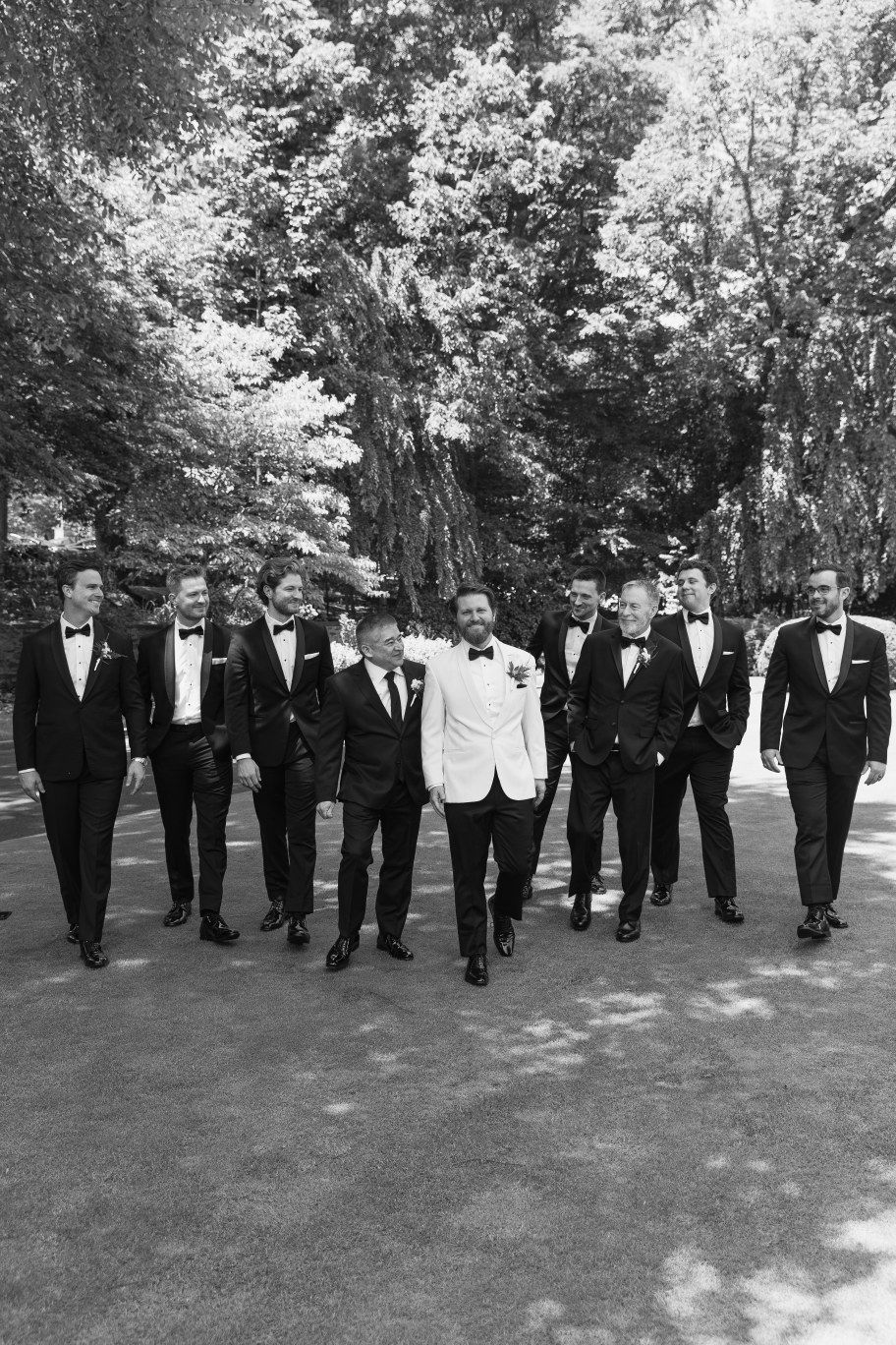 A group of men in formal black tuxedos and one in white walking together on a tree-lined path.