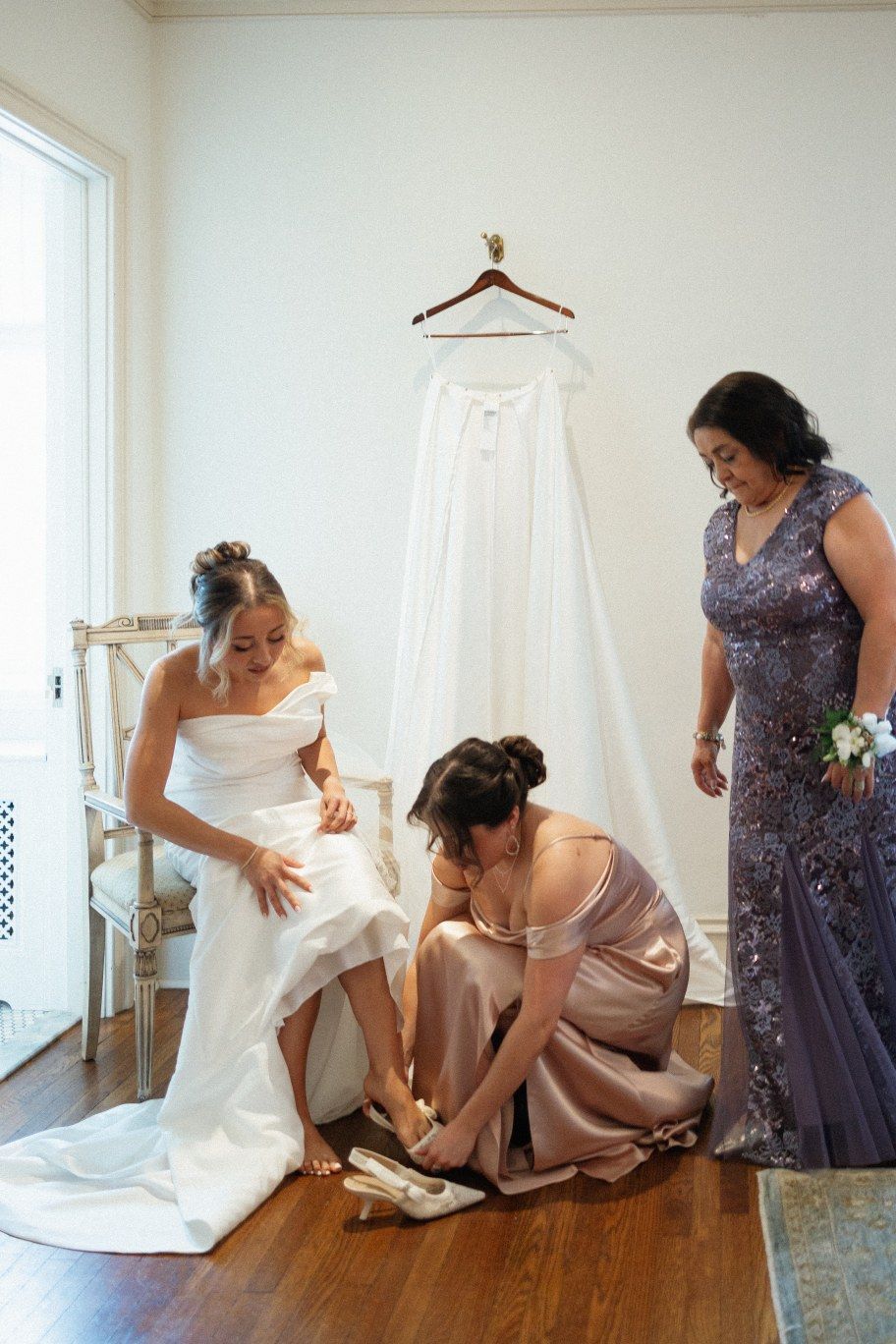 Wedding party helps bride get ready with her shoes while elegant white gown hangs on wooden hanger against wall.