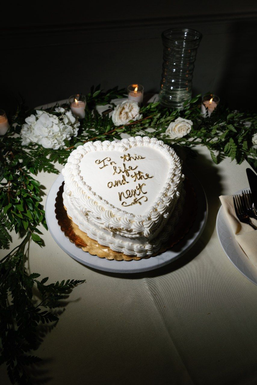 A white heart-shaped wedding cake with piped decorations sits on a table with greenery accents and candlelight.