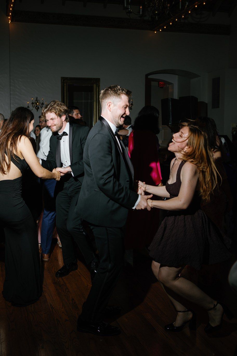 Couples dance together in formal attire at a dimly lit party with twinkling lights in the background.
