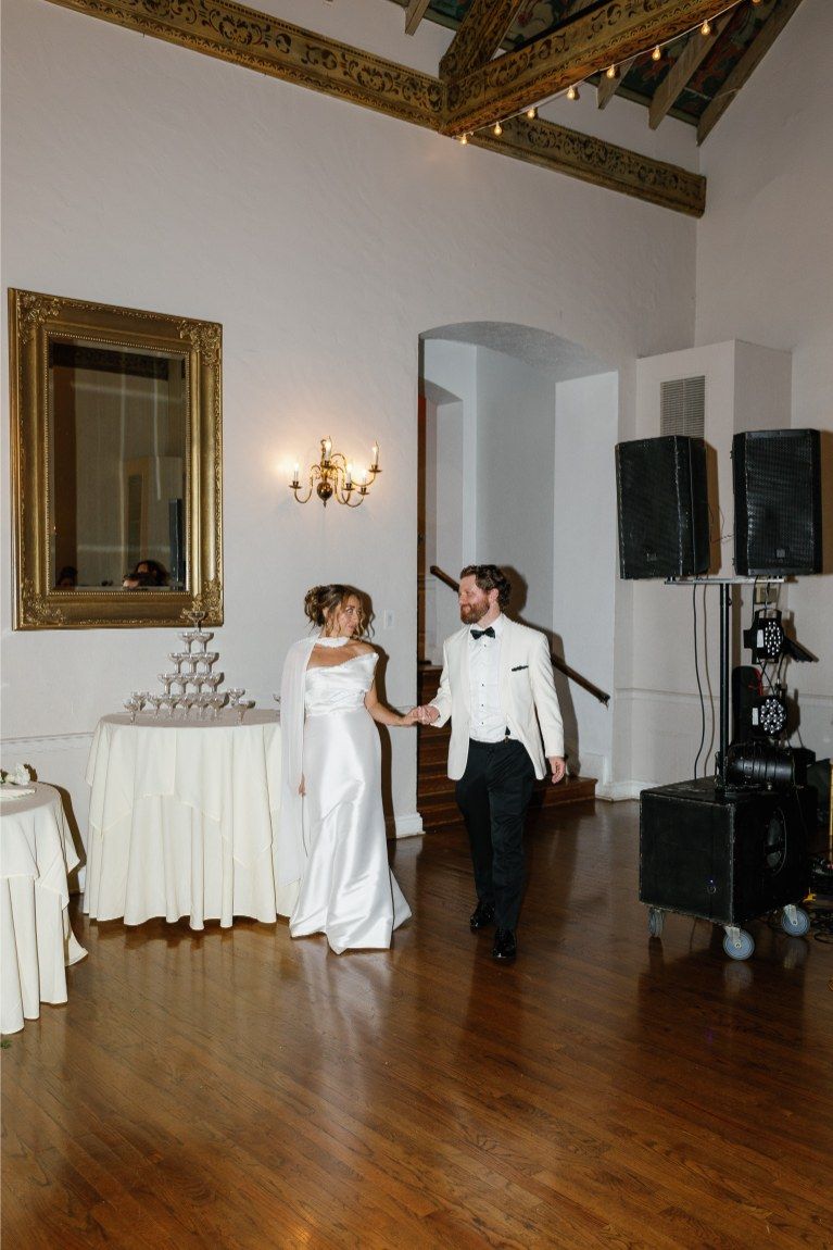 Empty banquet room with hardwood floors, ornate gold frames, and white-draped tables.