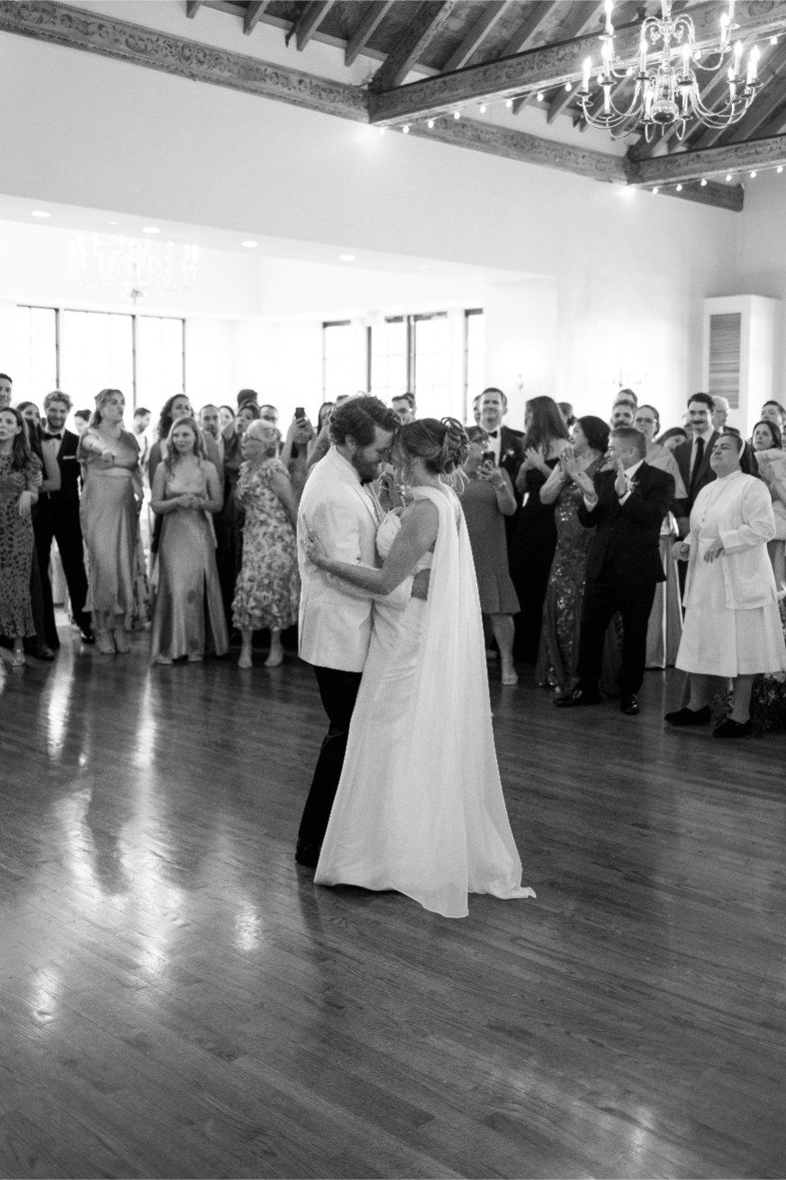 A newlywed couple shares their first dance surrounded by wedding guests in a grand ballroom with hardwood floors and chandeliers.