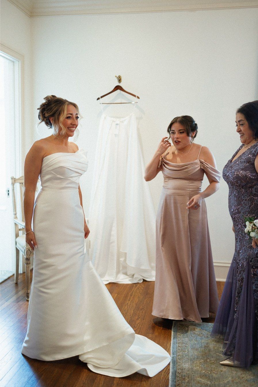 A joyful bridal scene showing three women in formal dresses standing in a room with a white wedding gown hanging on display.
