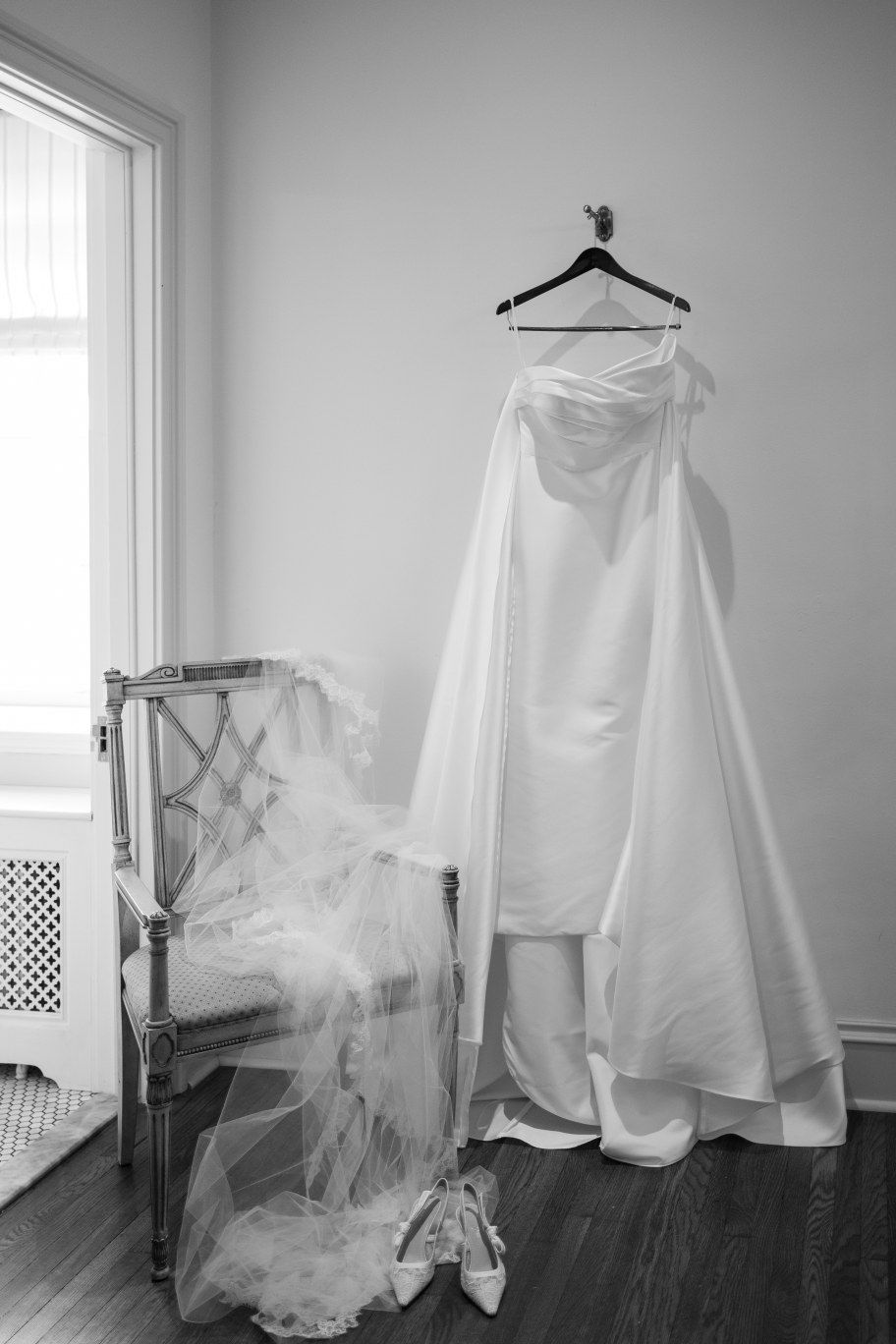 Black and white photograph of an elegant wedding dress hanging on a wooden hanger next to a chair with tulle veil.