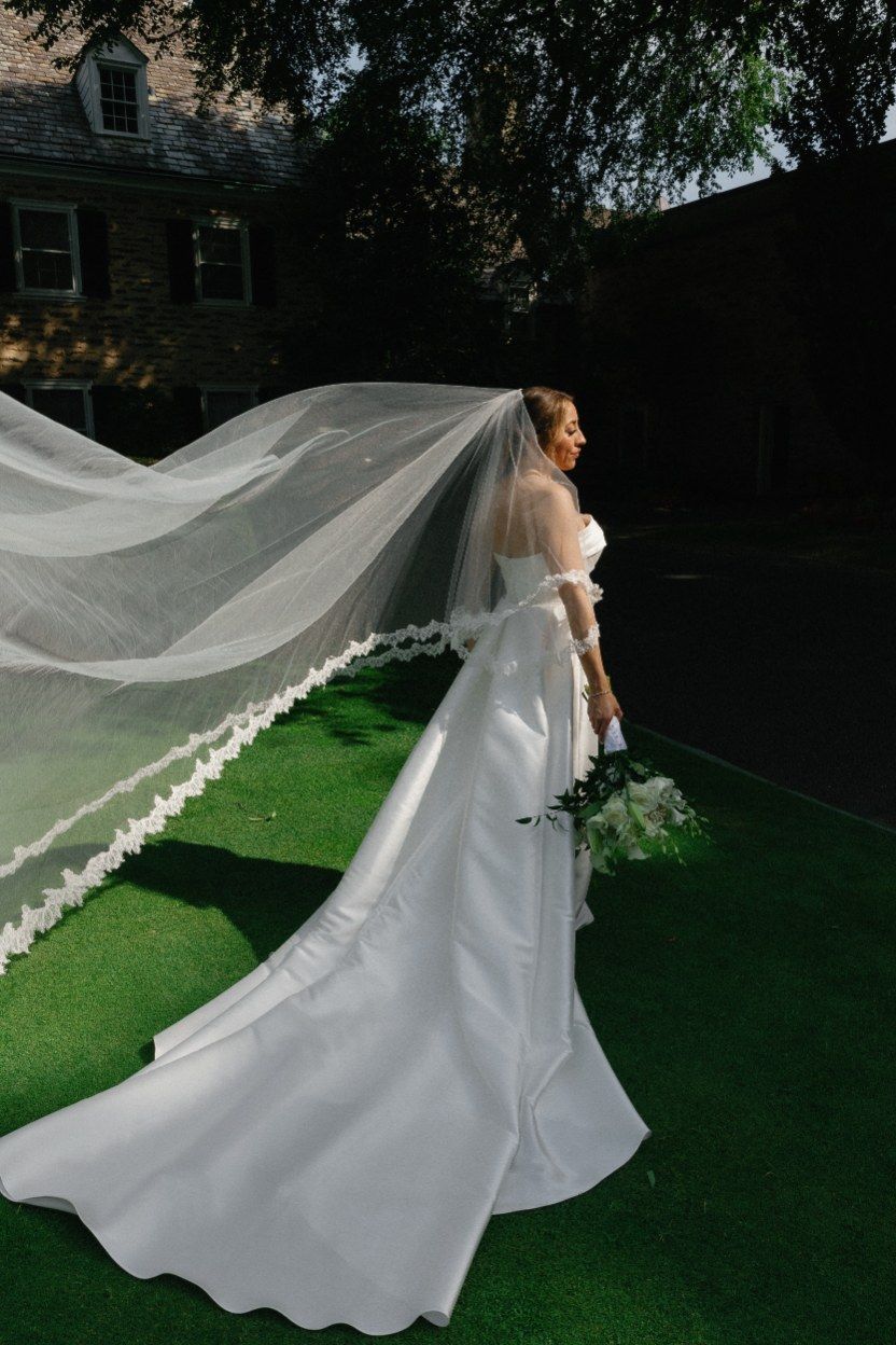 A stunning bride in a flowing white wedding dress with a dramatic long veil catches the wind in a garden setting.