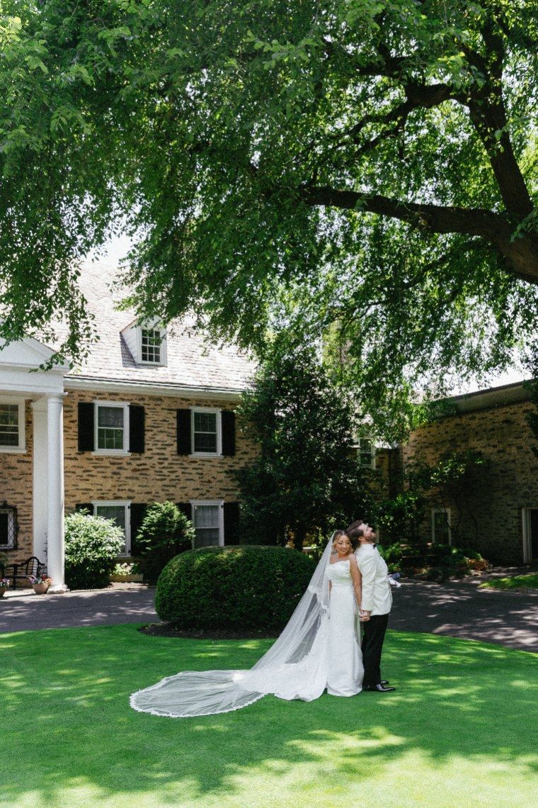 A couple shares a romantic moment on a lush lawn in front of a stone colonial house with white columns and overhanging trees.