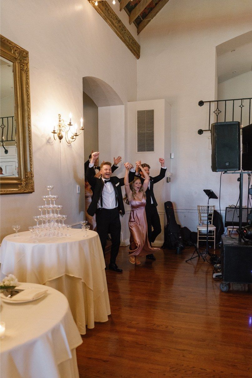 Two people enter an elegant reception room with hardwood floors, ornate mirror and a champagne tower display at a celebration.